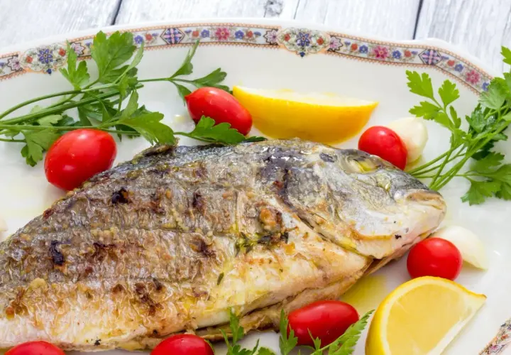 Orata alla griglia dorata e succulenta, guarnita con pomodorini, spicchi d'aglio, limone e prezzemolo fresco.