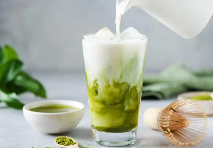 Milch wird in ein Glas mit Eis und Matcha gegossen. Ein Löffel mit Matcha-Pulver und eine Schale mit Matcha-Tee sind daneben zu sehen. Ein traditioneller Matcha-Besen liegt auf dem Tisch.