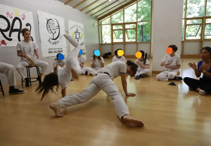 Bambini praticano capoeira in una palestra luminosa. Uno fa una capriola, altri osservano e applaudono.