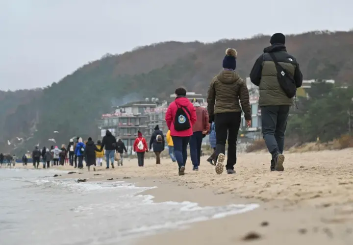 Spacer po plaży, gdy jest długi weekend. Ludzie w ciepłych ubraniach idą brzegiem morza, w tle widać zabudowania i wzgórza.