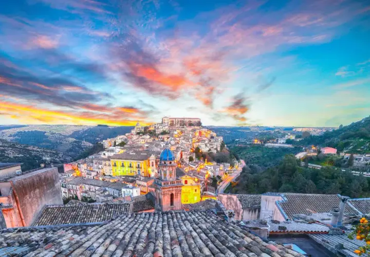 A picturesque Italian town perched on a hill, bathed in the warm glow of sunset. This view captures the essence of **two weeks in Sicily**, with its charming architecture and vibrant sky.