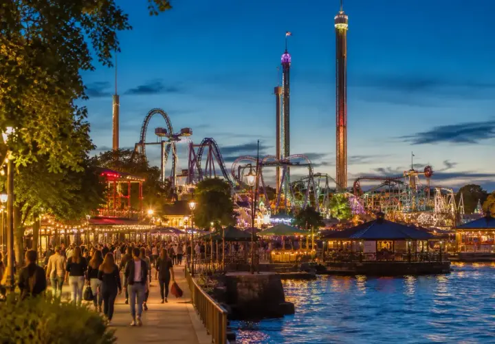 Kväll vid Gröna Lund med folk som promenerar längs vattnet, berg-och-dalbanor och höga torn som lyser upp himlen.