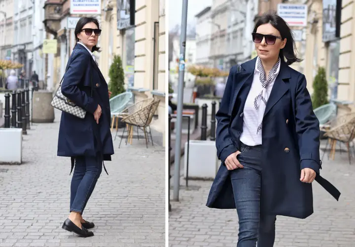 Elegancka kobieta w granatowym trenczu damskim, białej bluzce w paski i jeansach spaceruje po brukowanej ulicy.