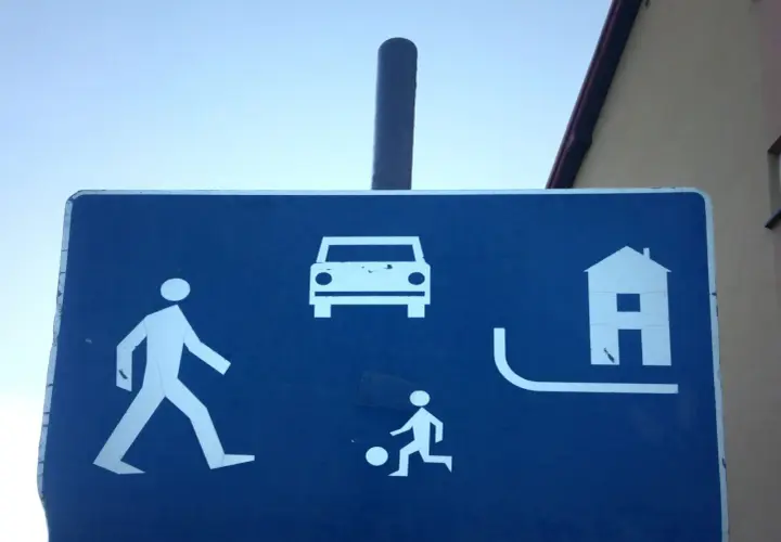 Znak informuje, że w strefie ruchu pieszy ma pierwszeństwo. Widać pieszego, dziecko grające w piłkę, samochód i dom.