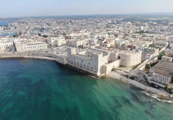 Vue aérienne du château de Trani, une forteresse médiévale sur la côte, avec la ville en arrière-plan et la mer d'un bleu-vert éclatant.
