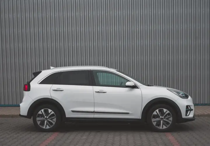 Biały Kia Niro EV stoi na tle szarej, falistej ściany. Elegancki design i nowoczesne linie podkreślają jego ekologiczny charakter.