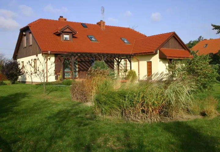 Dom z czerwonym dachem naczółkowym, z werandą i ogrodem pełnym traw ozdobnych.
