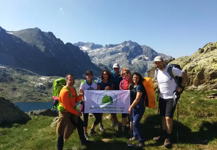 Grupo de excursionistas sonríe en la montaña, listos para la ruta. Llevan mochilas y un cartel que dice "Aquí empieza tu viaje".