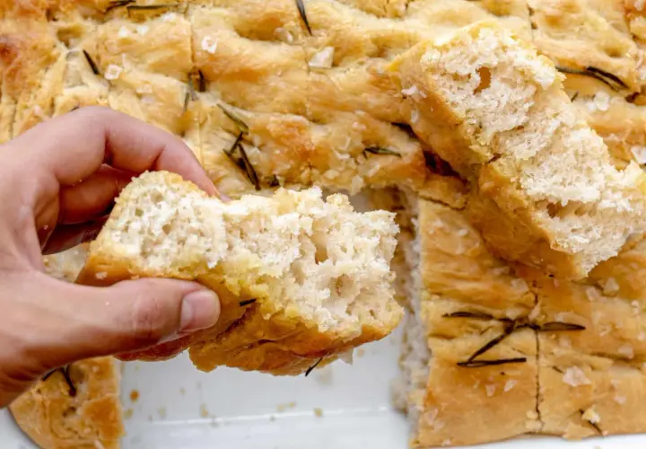 Mano rompe un trozo de focaccia original, dorada y con romero.