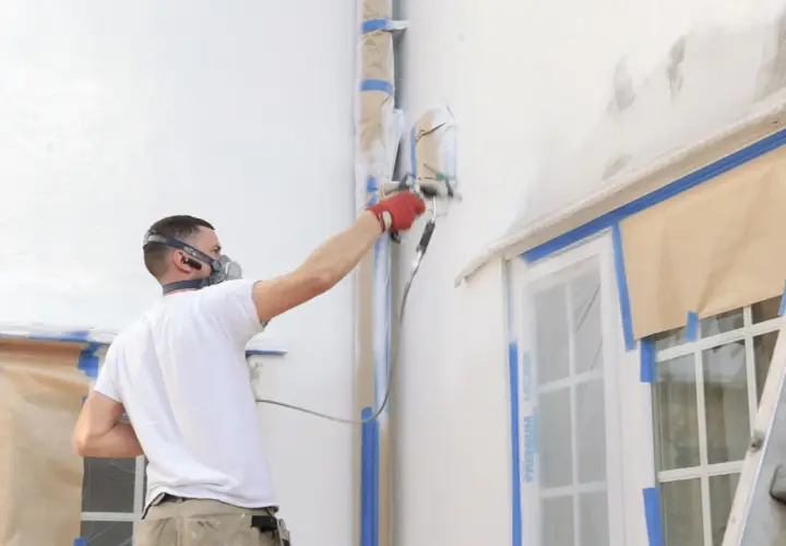 Uomo con maschera protettiva e guanti rossi esegue pittura a spruzzo su una facciata esterna, proteggendo finestre e tubi con nastro adesivo.