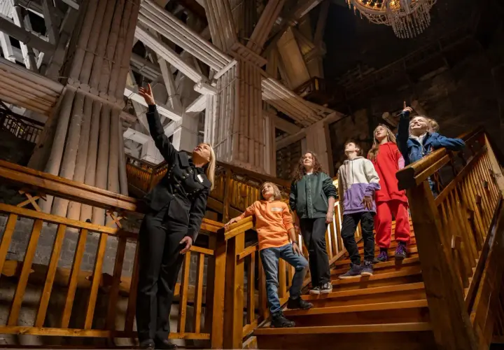 Przewodniczka oprowadza grupę dzieci po Kopalni Soli "Wieliczka". Wszyscy są ubrani na cebulkę, co sugeruje stałą, niską temperaturę w kopalni.