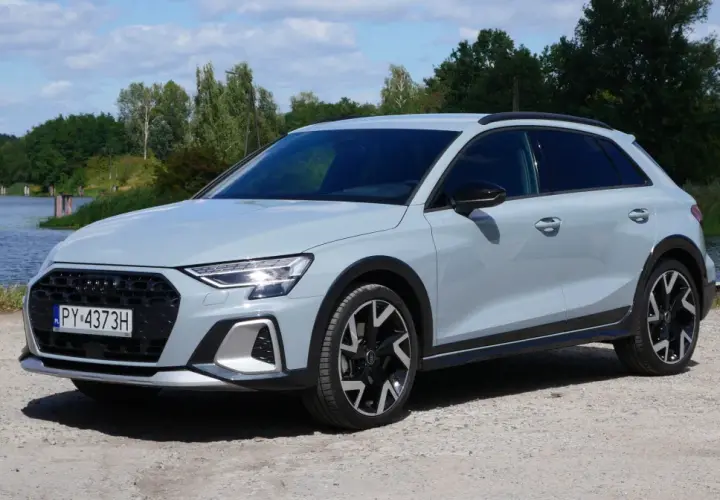 Jasnoszary Audi A3 Allstreet z czarnymi dodatkami stoi na żwirowej drodze nad jeziorem.