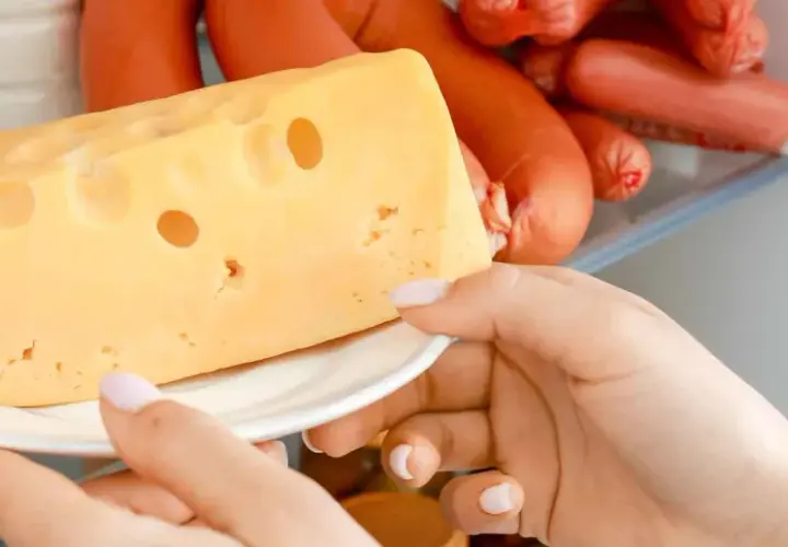 Una mano prende un pezzo di formaggio svizzero dal frigorifero, accanto a delle salsicce.