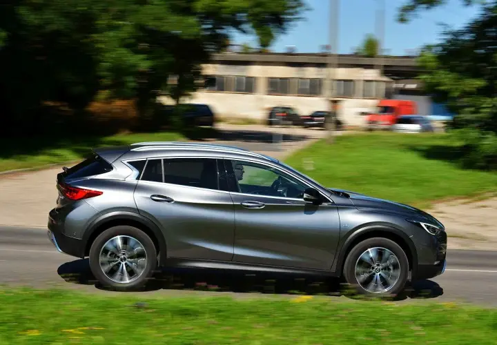 Srebrny Infiniti QX30 pędzi po drodze, mijając zielone pobocze i budynki.