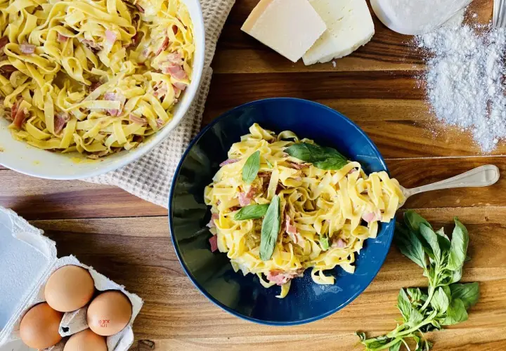 Un plat de pâtes à la carbonara crémeuses, garni de basilic frais. Des ingrédients comme des œufs, du fromage et de la farine sont visibles à proximité.