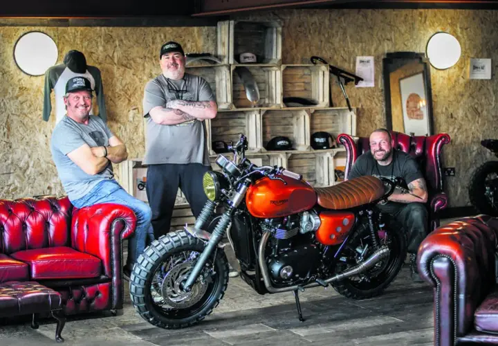 Three men pose with a custom orange Triumph motorcycle, showcasing the work of best custom bike builders.