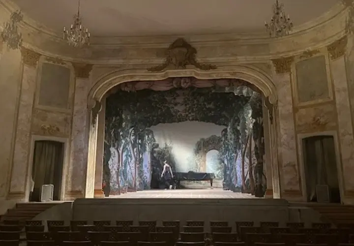 En ensam person vid ett piano på en scen med en skogsmålning. En vacker salong med kristallkronor, som påminner om **confidencen Ulriksdal**.