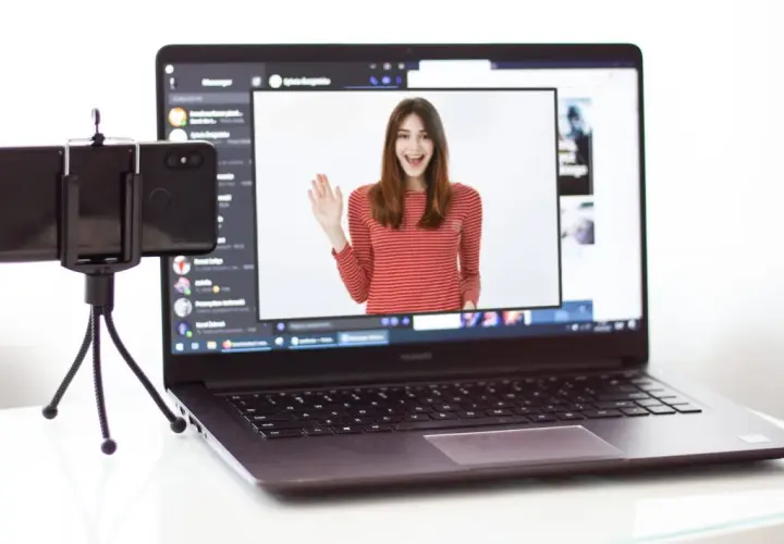 Kamera z telefonu na PC: młoda kobieta w bluzce w paski wita się na ekranie laptopa, obok stoi telefon na statywie.