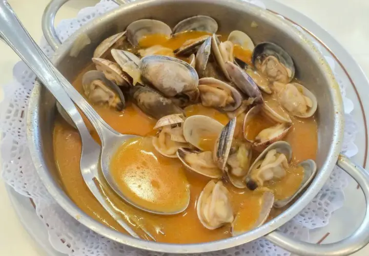 Almejas en una cazuela metálica, bañadas en una sabrosa salsa marinera. Una cuchara plateada reposa sobre el plato, lista para disfrutar.