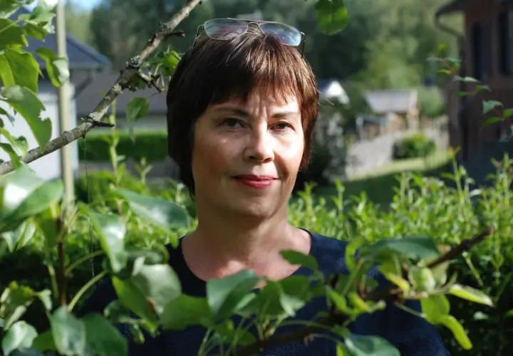 Anna Bratt, en kvinna med glasögon på huvudet, ler mot kameran i en lummig trädgård.