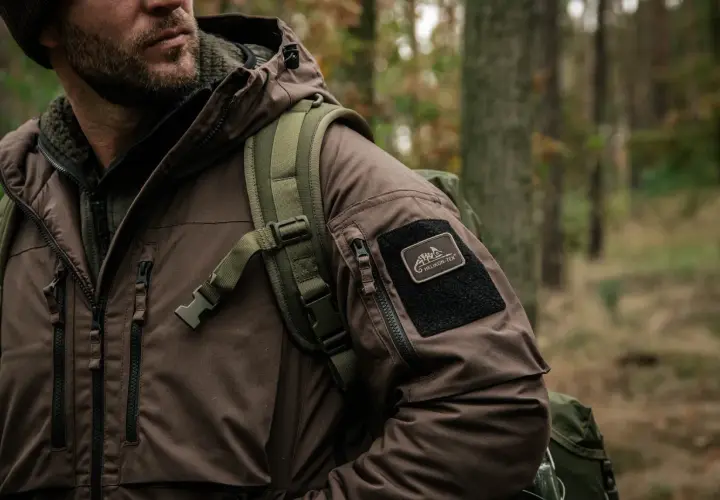 Mężczyzna w brązowej kurtce z naszywką Helikon-Tex, symbol polskie marki sportowe, w lesie.