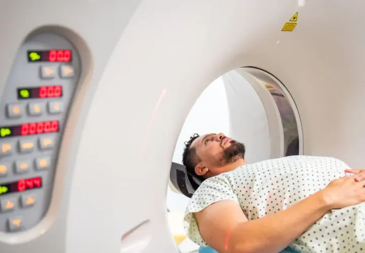 Mężczyzna leży w aparacie do tomografii komputerowej, przygotowując się do badania. Obok widoczny panel sterowania z cyfrowymi wskaźnikami.