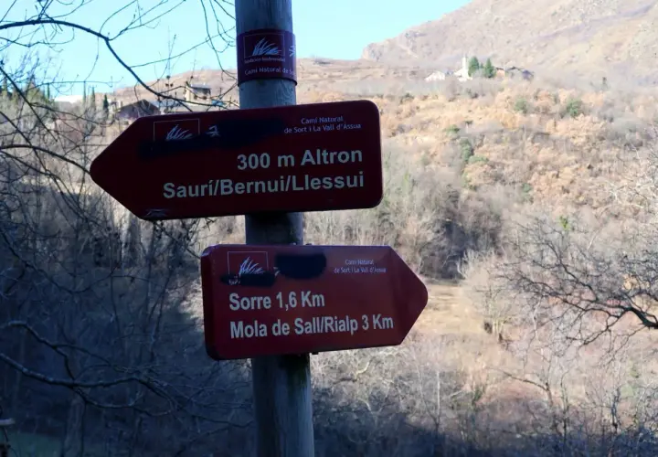 Señales de senderismo en la Vall d'Assua: 300m a Altron y 1,6km a Sorre. El paisaje montañoso y boscoso sugiere una ruta de naturaleza.