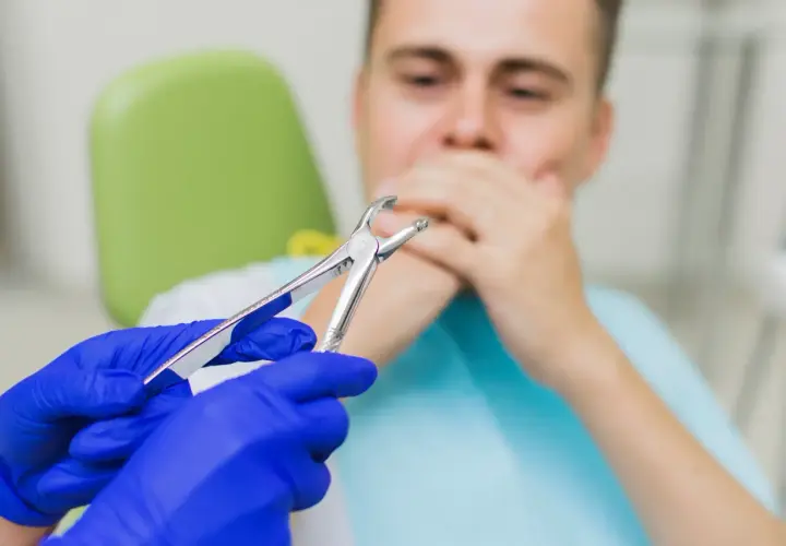 Dentysta w niebieskich rękawiczkach trzyma kleszcze. Pacjent zakrywa usta. Czy po wyrwaniu zęba można wychodzić na dwór?