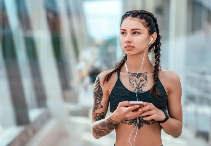 Kobieta z warkoczami i słuchawkami na uszach, trzymająca telefon. Widoczny tatuaż między piersiami przedstawiający kota.