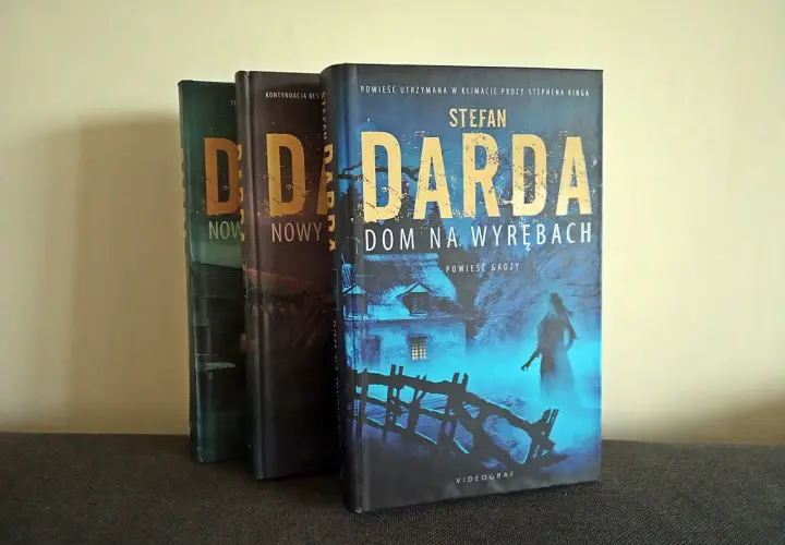 Trzy książki Stefana Dardy, w tym "Dom na wyrębach", stoją obok siebie. Okładka "Domu na wyrębach" przedstawia mroczny, niebieski krajobraz z domem i postacią.