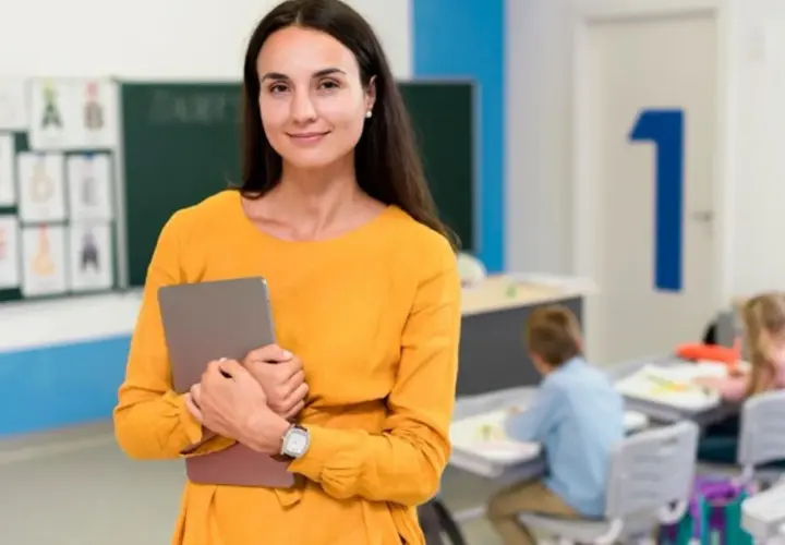 Nauczycielka w pomarańczowej sukience trzyma tablet w klasie, gdzie dzieci siedzą przy ławkach.