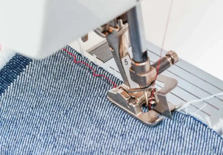 Sewing machine foot guiding denim fabric, demonstrating how to blind hem pants with a red thread stitch.
