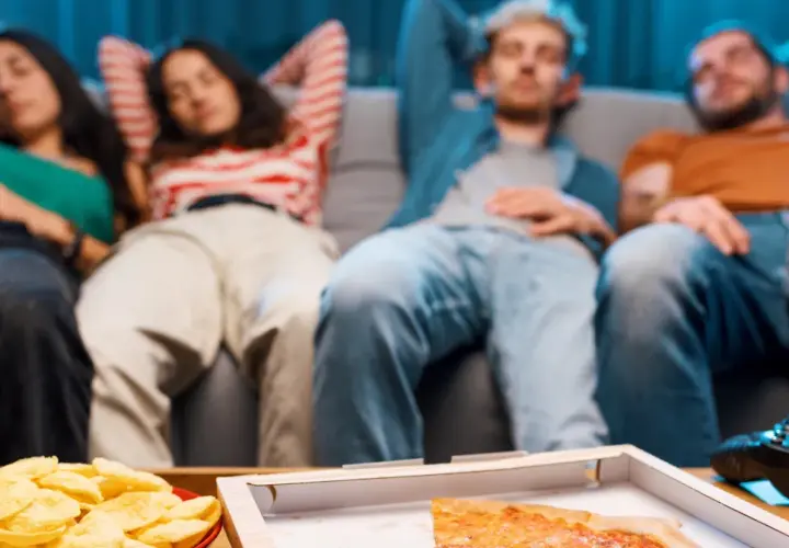 Grupa przyjaciół odpoczywa na kanapie po obfitym posiłku, czując senność. Na stole pizza, chipsy i piwo.