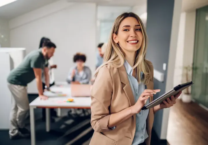 A smiling woman with a tablet, likely pursuing her masters in leadership and management, stands in an office as colleagues collaborate in the background.
