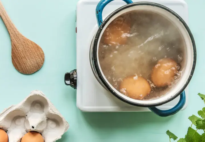 Des œufs sont en train de cuire des œufs durs dans une casserole d'eau bouillante. Une cuillère en bois et une boîte à œufs vide sont à proximité.