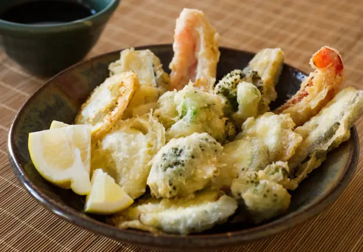 A bowl of crispy vegetable tempura, featuring broccoli, zucchini, and shrimp, served with lemon wedges and a side of dipping sauce.