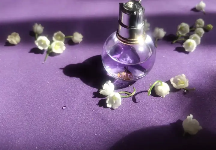 Szklana buteleczka perfum z fioletowym płynem i konwalią na fioletowym tle.