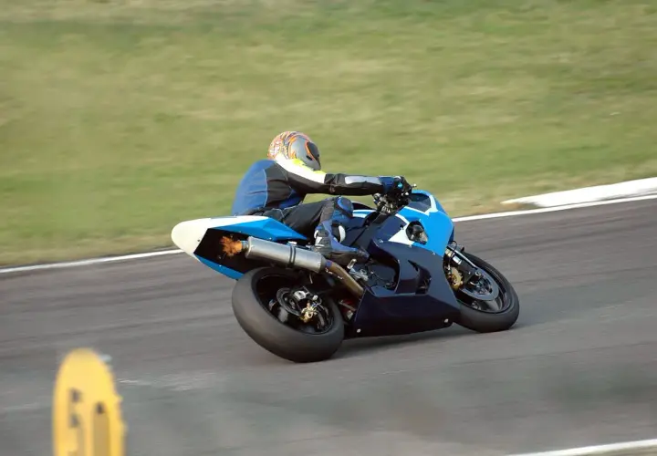 Piloto de MotoGP en plena curva, con la moto azul inclinada y llamas saliendo del escape. ¡Un auténtico holeshot!
