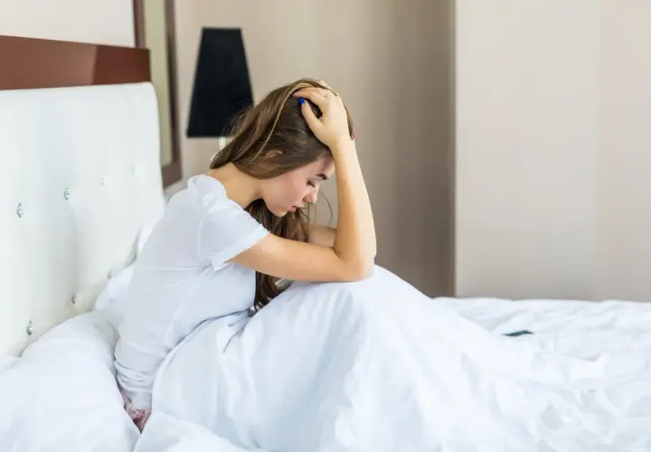 Mujer joven en la cama, con la cabeza entre las manos, sintiendo **ansiedad al despertar**.