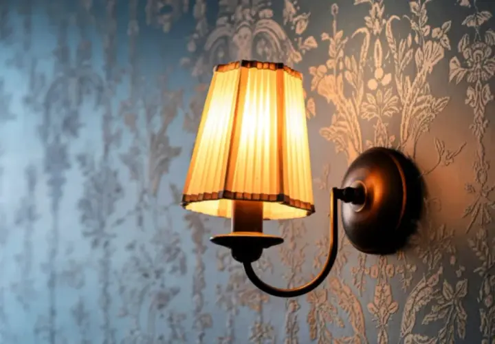 Aplique de pared rústico con pantalla plisada iluminada sobre papel tapiz floral.