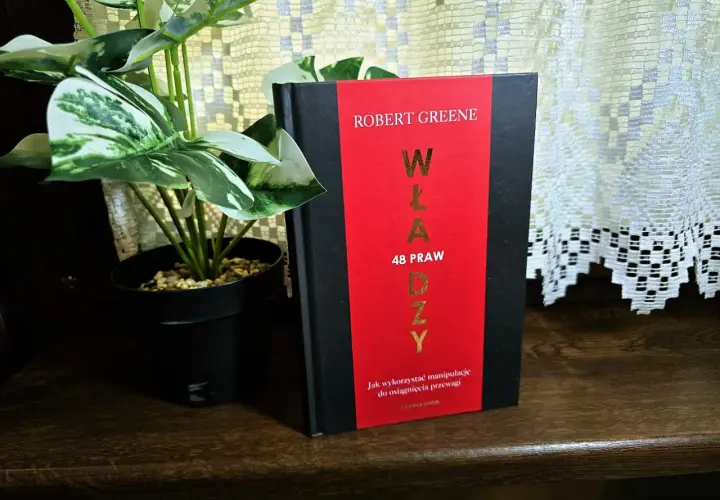 Książka "48 praw władzy" Roberta Greene'a na drewnianej półce, obok rośliny w czarnej doniczce.