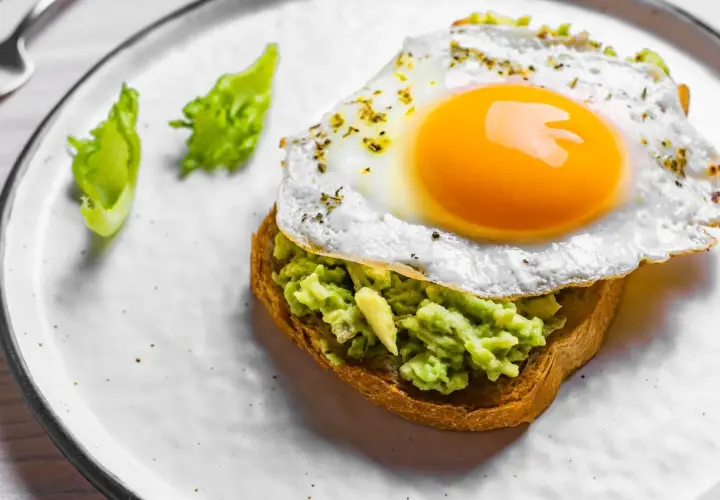 Un toast garni d'avocat écrasé et surmonté d'œufs sur le plat, assaisonné d'herbes. Deux feuilles de laitue verte décorent l'assiette.