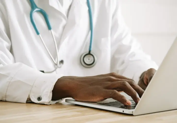 Mani di un medico che digita su un laptop, con uno stetoscopio appeso al camice bianco. Forse sta gestendo gli arretrati medici di base quando.