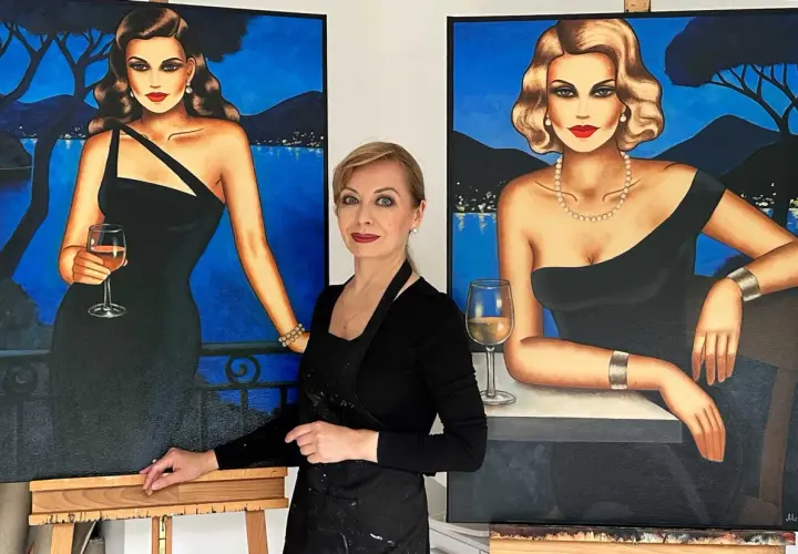 Künstlerin steht zwischen zwei Gemälden von eleganten Frauen in schwarzen Kleidern mit Gläsern Wein.
