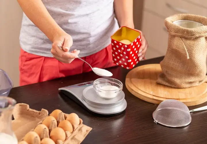Osoba odmierzająca cukier łyżką na wagę kuchenną, obok jajka i mąka w worku.
