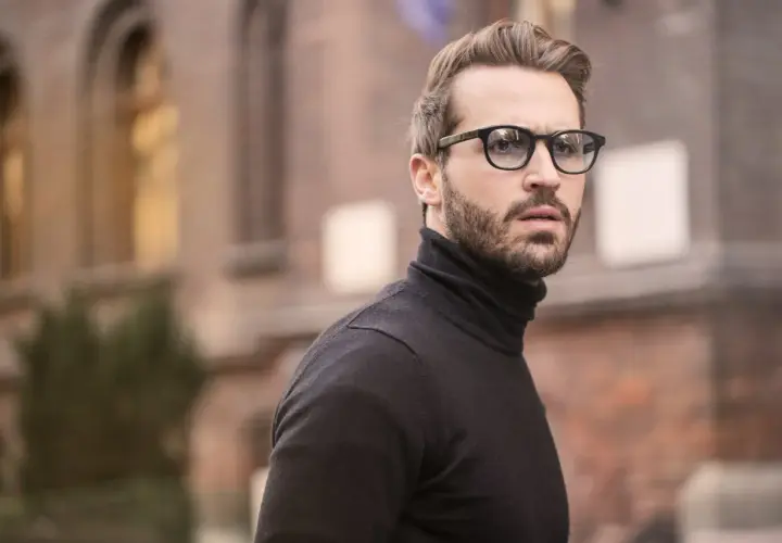 Hombre con barba y gafas, luciendo un jersey negro de cuello alto. Un estilo moderno que realza los rasgos, ideal para cortes para cara redonda hombre.