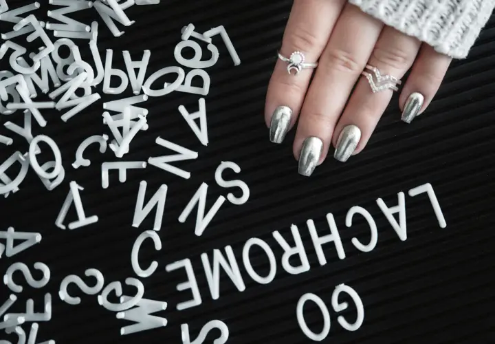 A hand with silver metallic nails and rings rests on a black surface scattered with white letters.