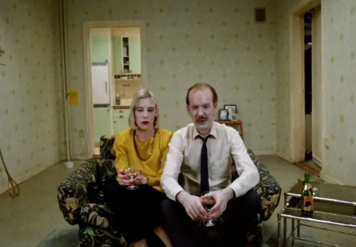 Ett par sitter i en soffa, en kvinna med gul tröja och en man i skjorta och slips. Kanske en scen från en Roy Andersson reklamfilm.