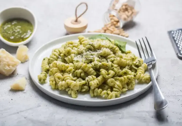 Pyszny makaron z pesto ze słoika, posypany parmezanem. Obok miseczka z zielonym sosem i kawałki sera.