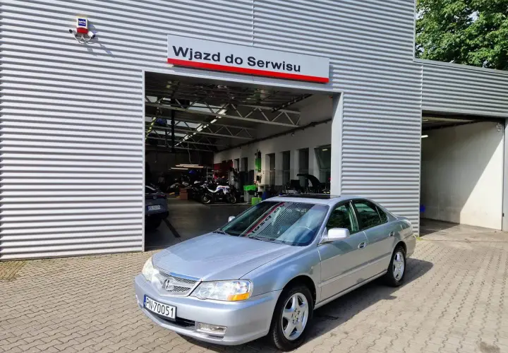 Srebrny sedan przed wjazdem do serwisu. W tle widać motocykle i sprzęt warsztatowy. Może to miejsce, gdzie naprawią mój carprog2?
